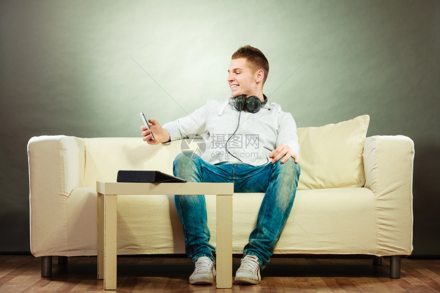 年轻英俊男子在沙发上放松家用耳机智能手和平板电脑图片
