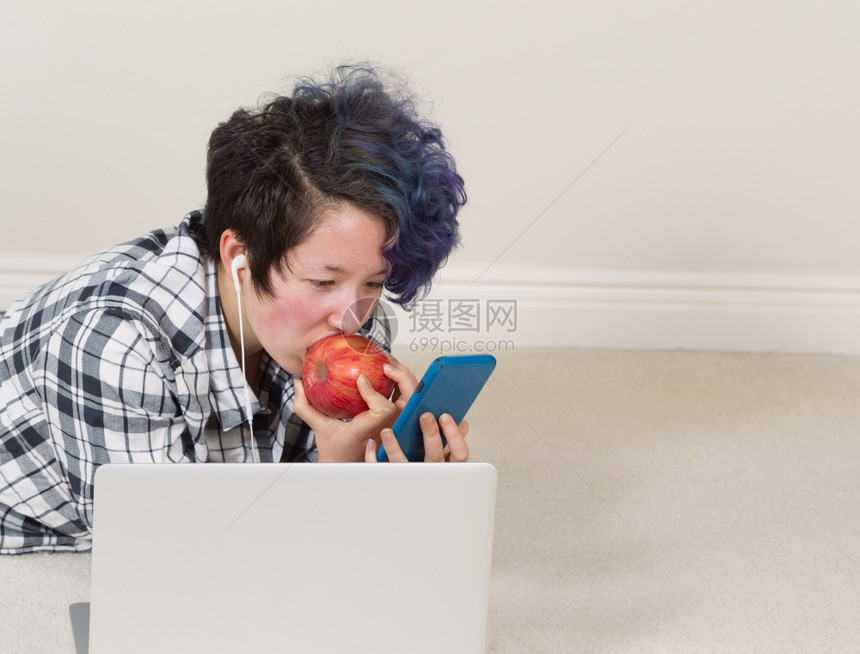 女孩一边吃苹果看手机前面有电脑一边躺着听家里的音乐