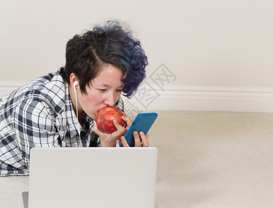 女孩一边吃苹果看手机前面有电脑一边躺着听家里的音乐青少年高清图片素材