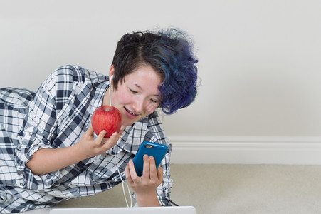年轻女孩拿着苹果一边看手机前面有电脑一边躺着听家里的音乐地板高清图片素材