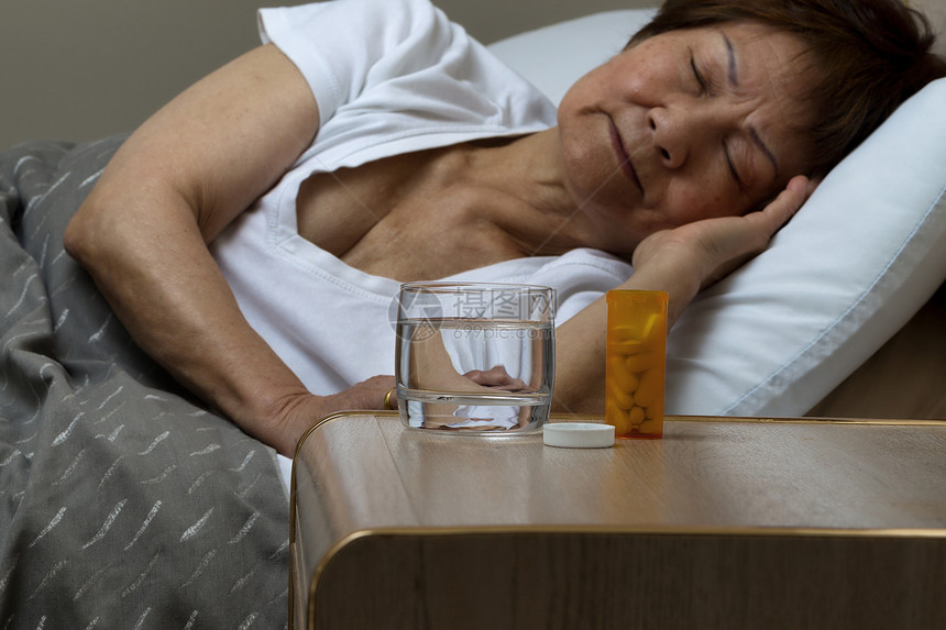 睡前柜上紧贴着一瓶开的药和一杯水睡在背景中的老年妇女睡在床头柜上图片