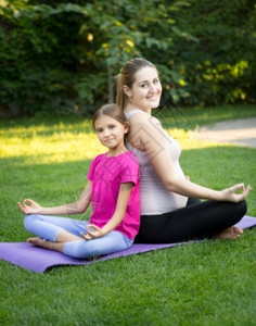 快乐的母亲和微笑女儿做瑜伽坐在公园后着图片