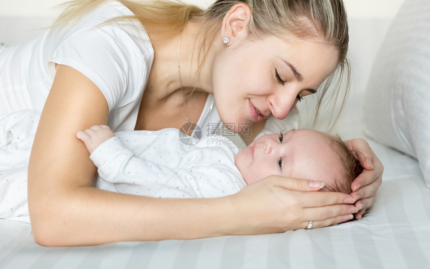 美丽年轻母亲抱着婴儿男孩躺在床上的近身肖像图片