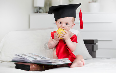 从书本和咬苹果上坐着的毕业帽子上可爱的小男孩肖像图片
