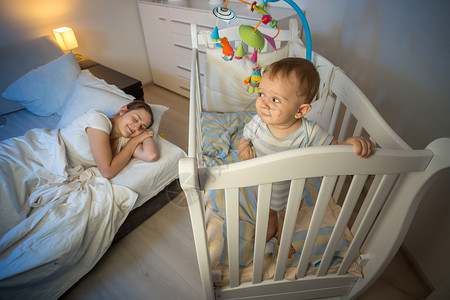 可爱宝的肖像站在婴儿床看着睡的疲累母亲图片