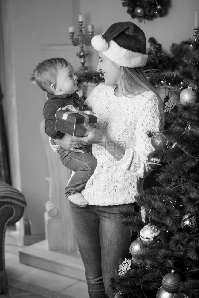圣诞老人帽子中快乐的年轻母亲和10个月大的婴儿子在客厅圣诞树上摆着的画像图片