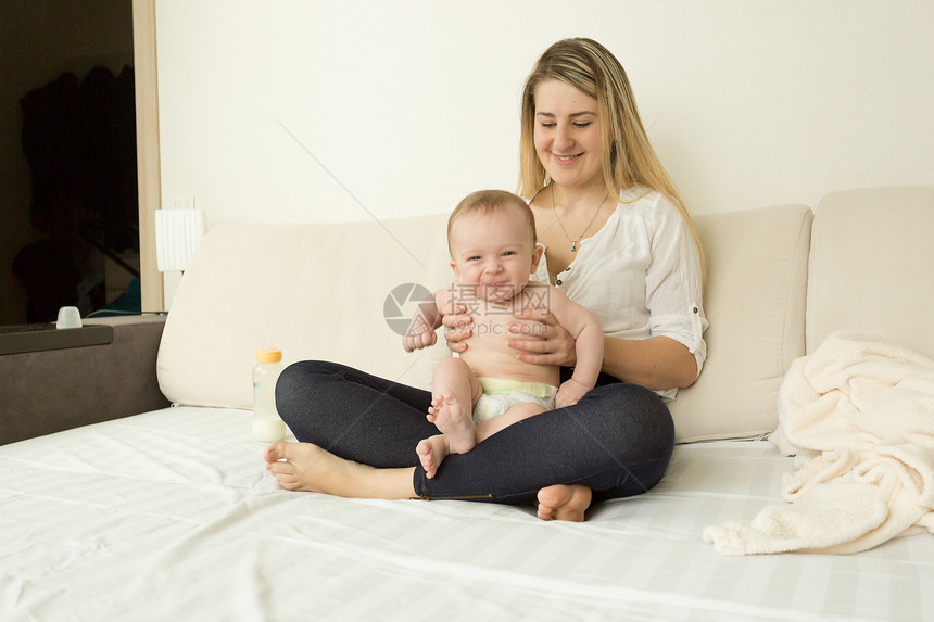 微笑的年轻母亲与婴儿男孩坐在床上图片