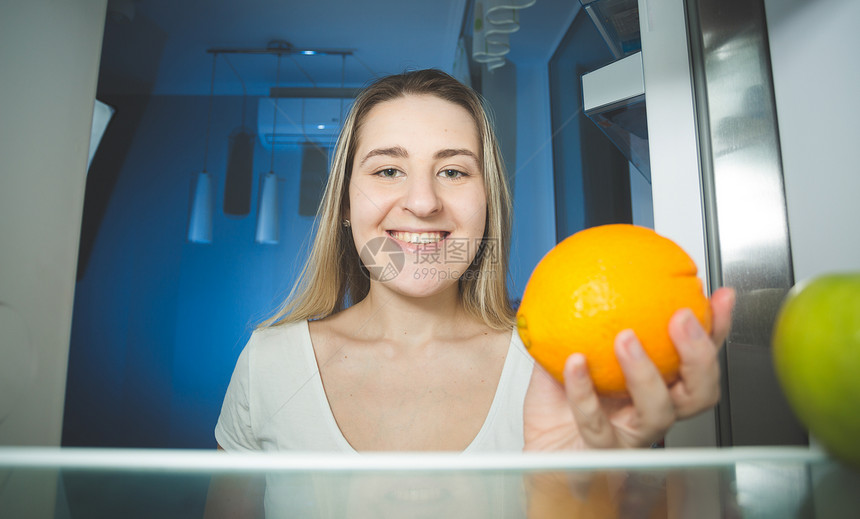 美丽的微笑女人拿着橙色看冰箱里图片