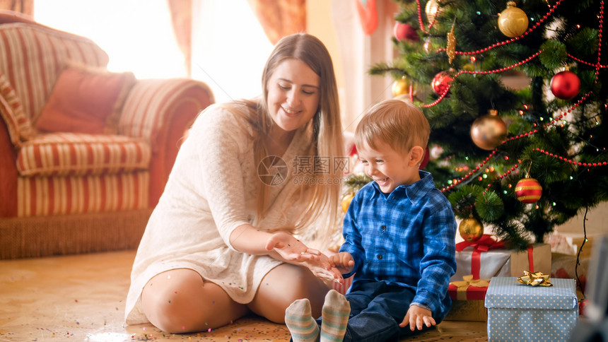 欢笑的小孩男与美丽的年轻母亲躺在地板上面盖着圣诞树旁边的多彩蛋白欢笑的小孩男与美丽的年轻母亲躺在地板上面盖着多彩蛋白在圣诞树旁边图片