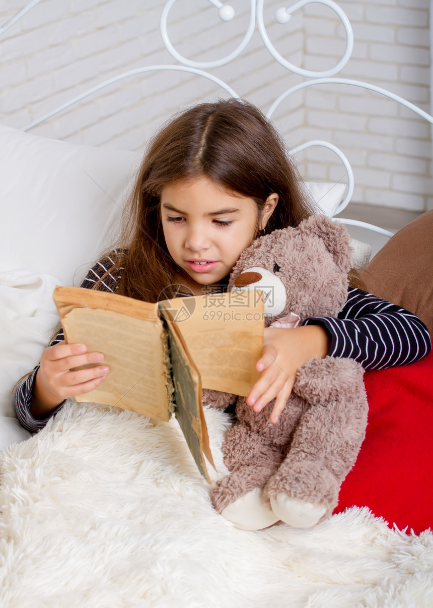 小女孩躺在床上和她最喜欢的泰迪熊躺在床上睡觉前读一本书图片