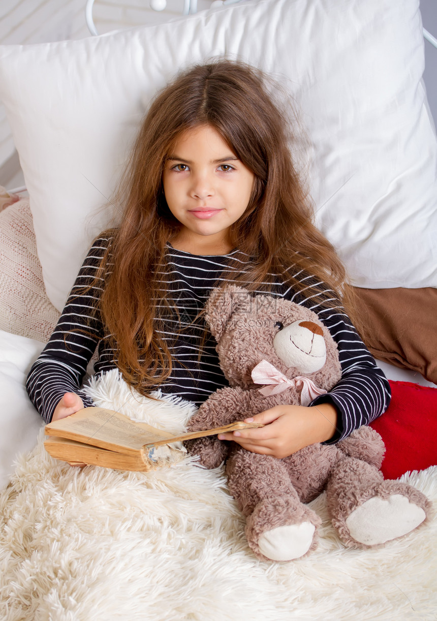 小女孩躺在床上和她最喜欢的泰迪熊躺在床上睡觉前读一本书图片