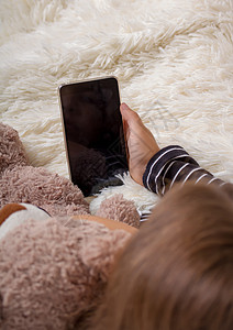 小女孩躺在床上带着她最喜欢的泰迪熊躺在床上玩智能手机看高清图片素材