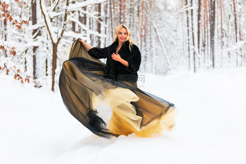 穿着长的黑黄色金裙子的发美女在雪的冬季森林或户外的木中图片