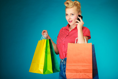 Pinup金发女孩年轻旧式呼叫手机客户持有多彩的纸袋生机勃的蓝色零售和销工作室拍摄背景图片