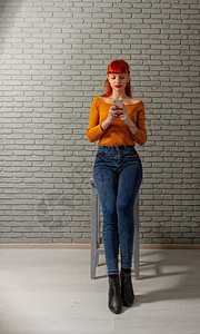 年轻红头发女孩在她的智能手机上用灰砖墙对着看东西图片