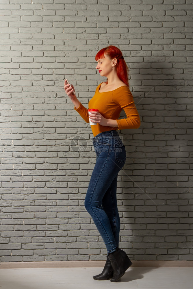 年轻红头发女孩带着一杯咖啡正在她的智能手机上读到一些深思熟虑的东西图片