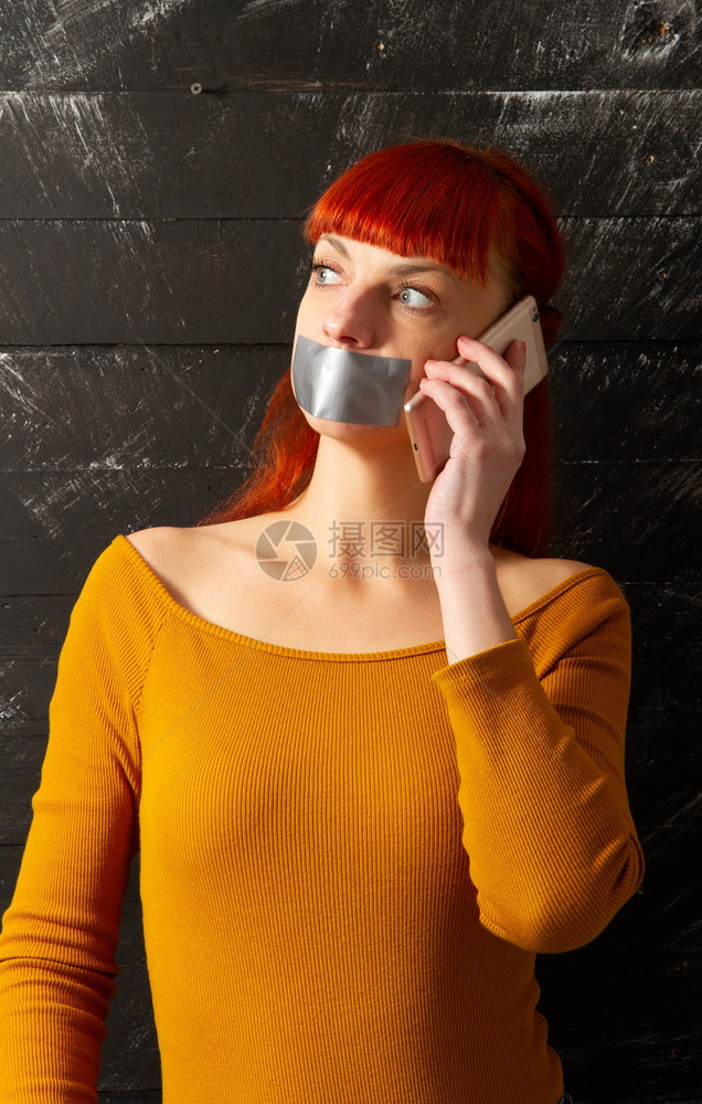 年轻红头发女孩试图在电话上说用粘合胶带封住用嘴唇以便保持缄默图片