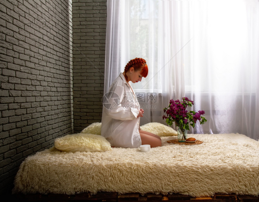 一名年轻红头发女孩早上坐在窗户附近的低层床上只穿着男子和穿浅色衬衫喝咖啡图片