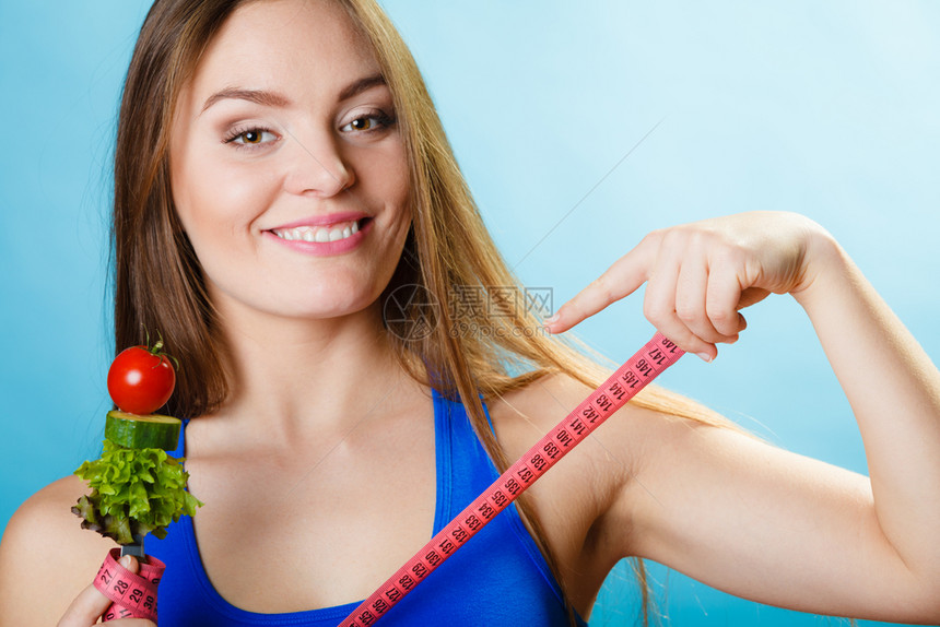 健美的身女拿着新鲜混合蔬菜的叉子和蓝色背景的测量胶带图片