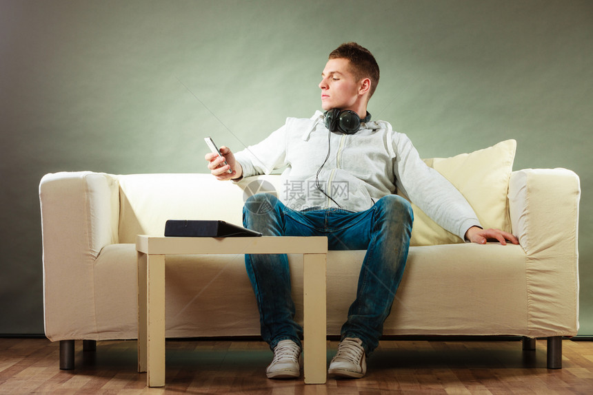 年轻英俊男子在沙发上放松家用耳机智能手和平板电脑图片