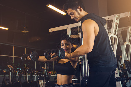 个人训练男子帮助妇女手臂拉起哑铃在室内健身房锻炼图片