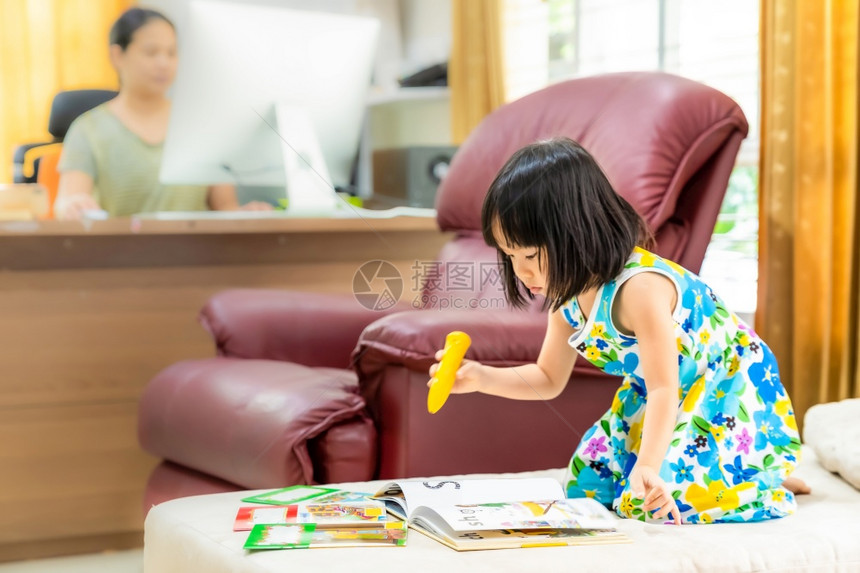 亚洲女童在家里客厅阅读互动式书籍作为家庭教育而她的母亲则在家里工作因为城市由于新冠19大流行病而被封锁图片