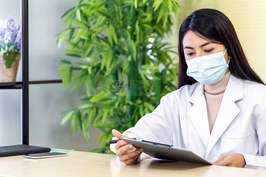 Asian女医生的肖像是戴保护面罩的亚洲女医生坐在院诊所的办公室里在检查耐心之前先阅读文件然后检查耐心图片