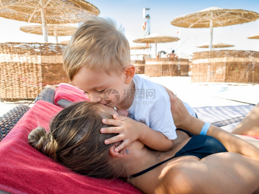 3岁可爱的男孩肖像亲吻他母在阳光保护伞下海边放松家庭在暑假期间沙滩上放松和愉快3岁可爱的幼儿男孩肖像在阳光保护伞下海边放松图片