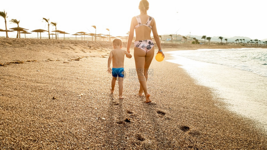 快乐的年轻母亲用手抱着她小孩的美丽形象在寒冷的海滨上行走与惊人的日落天空相对的温暖平静波浪相对美丽的年轻母亲用手抱着她小孩的美丽图片
