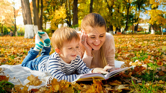 快乐微笑的男孩和母亲秋天在公园阅读图片