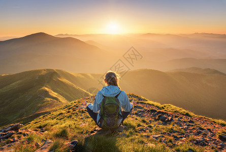 穿着背包坐在山峰上的年轻女子和美丽的山峰在夏季日落时雾中与运动女郎绿林山丘天空太阳光旅行和游瑜伽背景图片