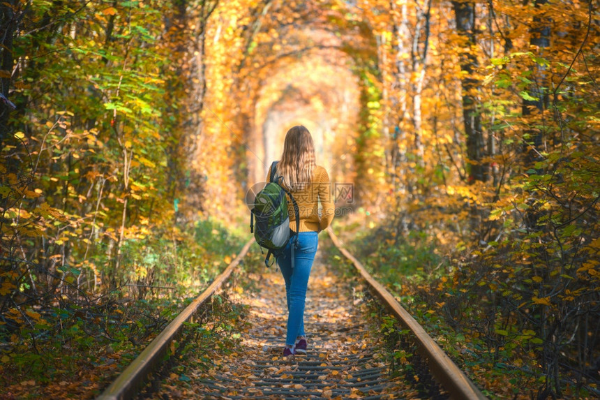 秋天日落时在乌克兰爱隧道的铁路上行走年轻女子在火车站上背着包的女孩在多彩树木的隧道里倒下美丽的森林中铁路生活方式秋天在铁路上行走图片