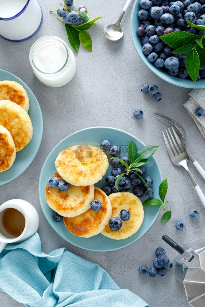 奶油芝士煎饼甜卷和浆果配蜜蜂新鲜蓝莓的西尼基饼和早餐桌上的新鲜蓝莓图片