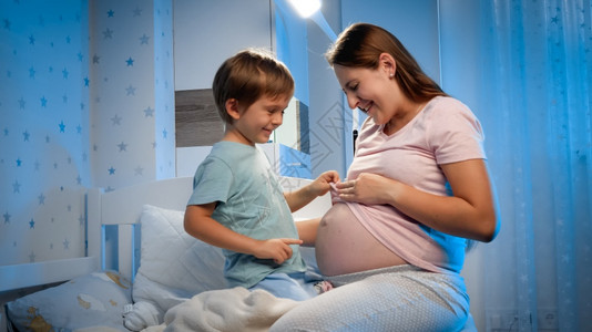 Smilk幼儿男孩看着和摸他怀孕母亲的大肚子晚上在床等婴儿图片