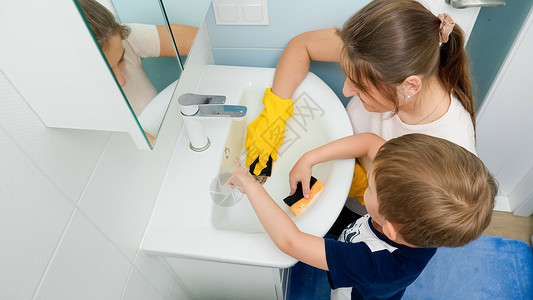 快乐的男孩母亲戴着橡皮手套在海绵上涂洗涤剂和清洁洗浴室水池小男孩帮助父母做家务快乐的男孩在海绵上涂洗涤剂和浴室水池背景图片