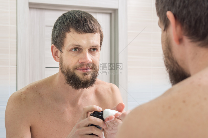一个未剃毛的人用须泡沫在他的手掌上看着在浴室的镜子图片