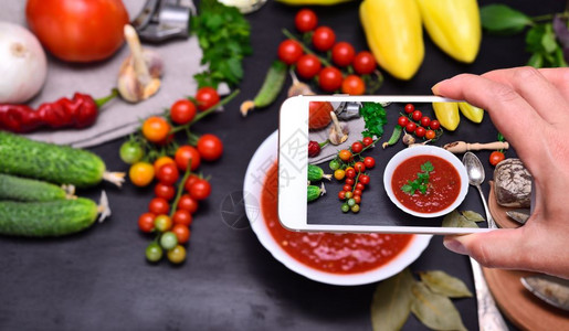 女手拿智能手机拍摄白色圆形盘子里红色西红柿汤的gazpacho俯视图图片