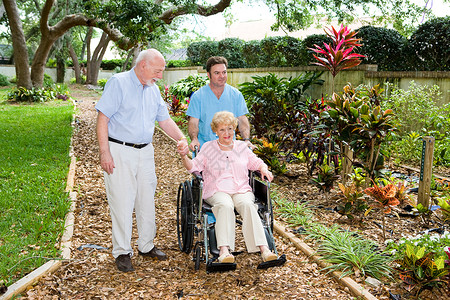 坐在轮椅上的年长妇女正由一名修士及其丈夫步行穿过疗养院花园手高清图片素材