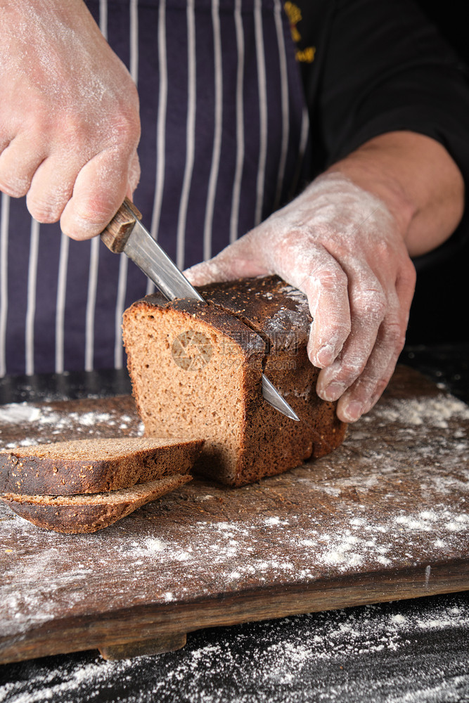 身穿黑色制服的厨师手里拿着一把厨房刀在木板上切掉烤黑麦面粉包上的片图片
