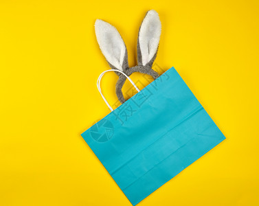 黄色背景的白手柄长方形蓝色纸购物袋中露出兔子耳朵平躺着图片