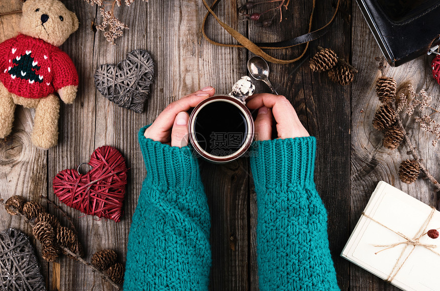 妇女手握着一件绿色编织的毛衣手持一个红陶瓷杯黑咖啡灰色木桌图片