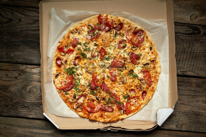 用熏香肠蘑菇西红柿奶酪和薄饼烤圆披萨在木制餐桌上的开板纸盒中食物被切成片顶视图片