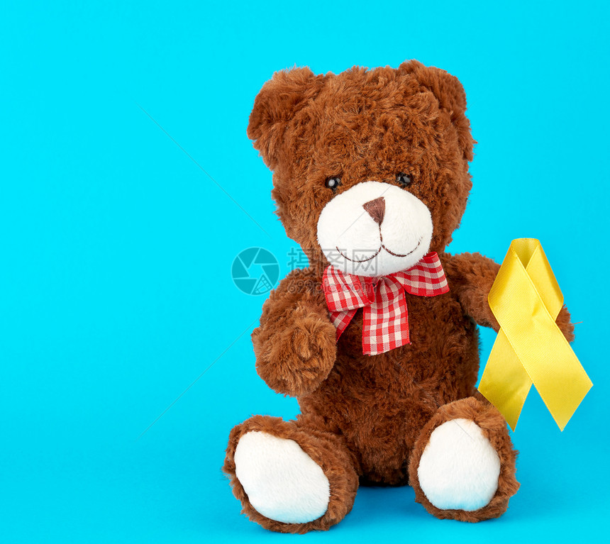 棕色泰迪熊坐在他的爪子上,持有蓝色背景的黄丝带,与儿童癌症作斗争的概念。 问题及其预防。图片