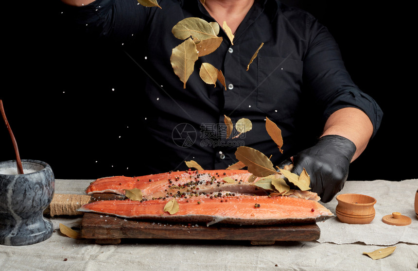 穿黑衬衫的成年男子倒白粗咸盐和干叶放在新鲜鲑鱼肉片烹饪过程黑暗背景上图片