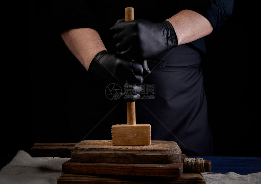 穿黑色制服的男厨师拿着木锤子用黑色背景的旧餐板打肉图片