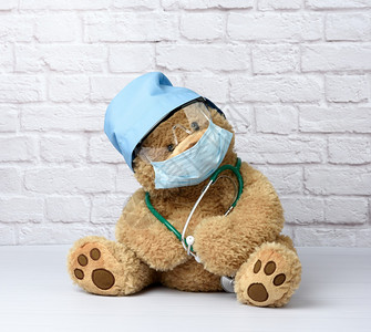 棕色泰迪熊坐在保护塑料眼镜医用一次面罩和白色砖墙背景下的蓝帽图片