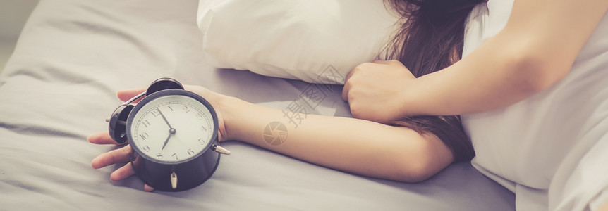早上关闭闹钟醒来睡觉时女孩拿着闹钟时间和生活方式的概念图片