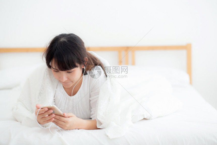 美丽的年轻女躺着快乐的耳机在线听音乐放松和享受在卧室女孩玩耳机歌曲智能手机生活方式休闲概念图片