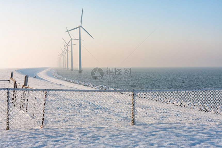 在冬季荷兰在河中与雪栅栏和长排风力涡轮机在冬季烟雾中缺水的离岸停靠长排风力涡轮机一起冬荷兰在季用雪围栏和风力涡轮机起水图片