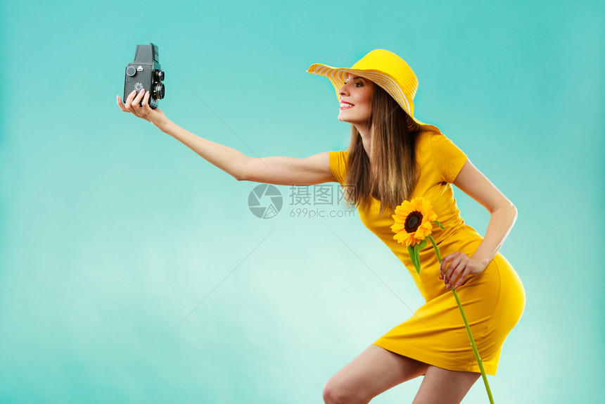 身着黄色洋装和向日葵帽子的暑期妇女穿着黄色洋装戴向日葵帽用蓝背景的旧照相机自拍图片